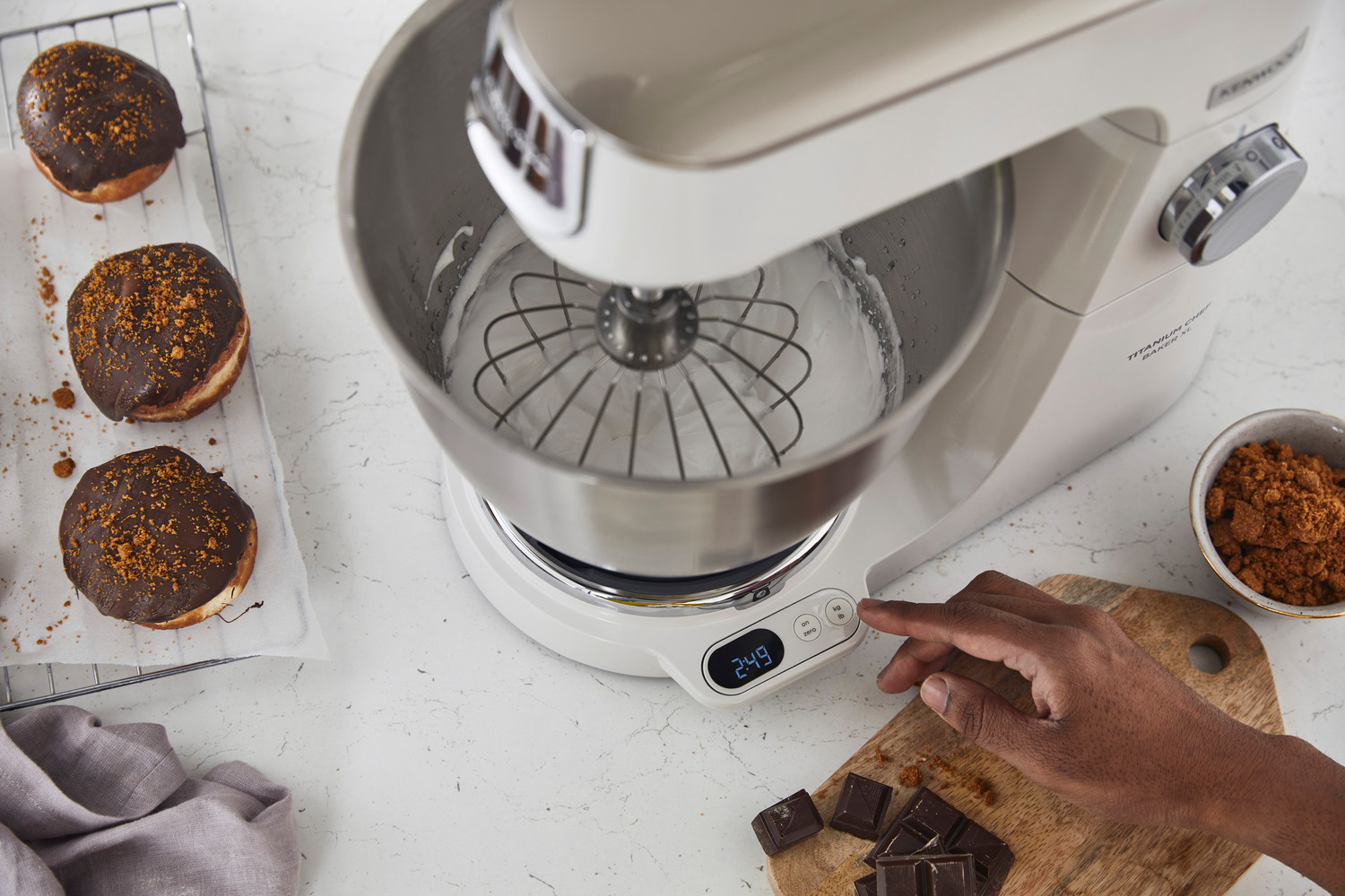 Weißer Küchenmixer mit Schneebesenaufsatz. Donuts und Schokolade an der Seite.