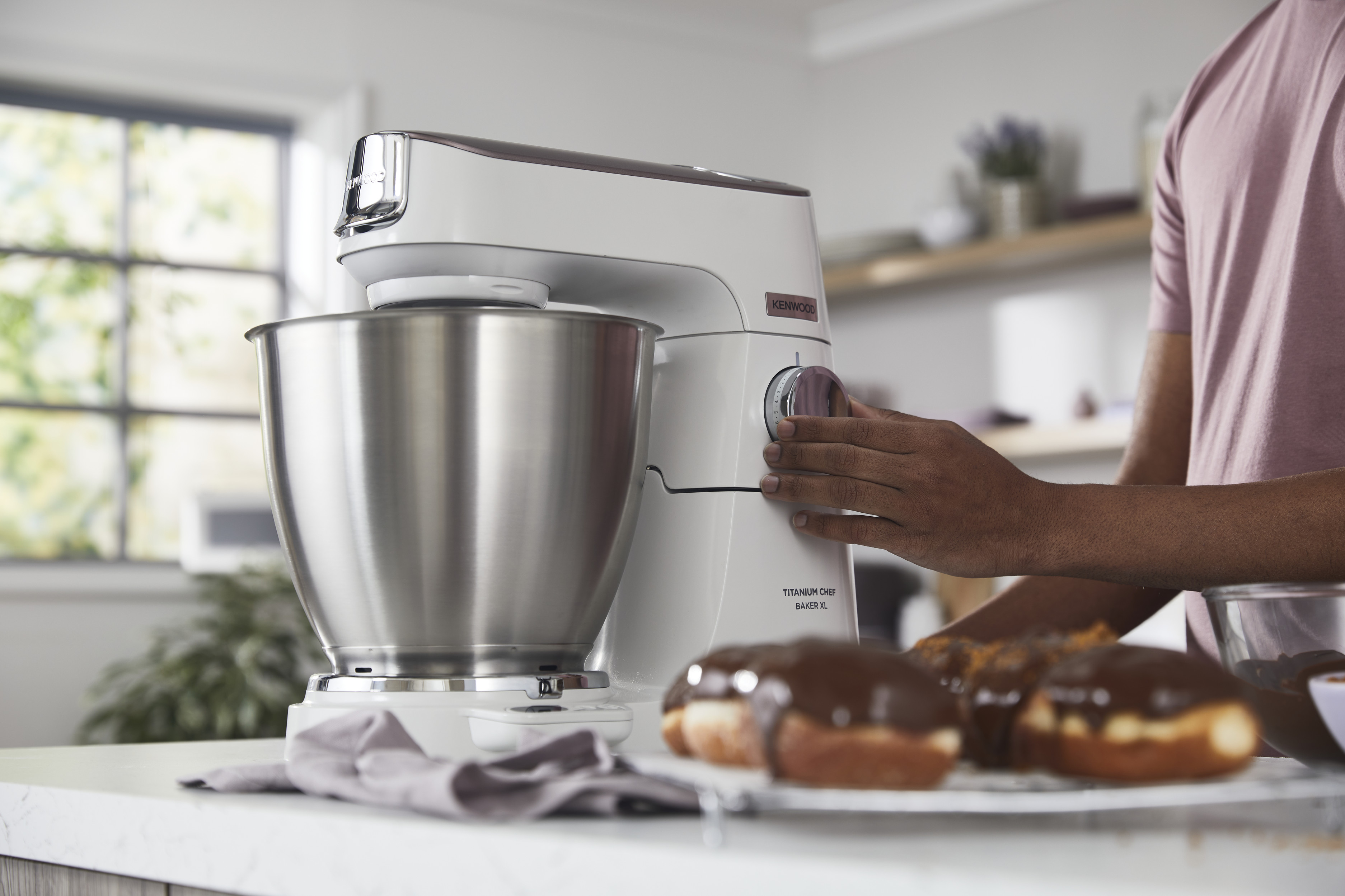 Eine Person, die einen weißen Kenwood Küchenmixer benutzt. Donuts mit Schokolade daneben.