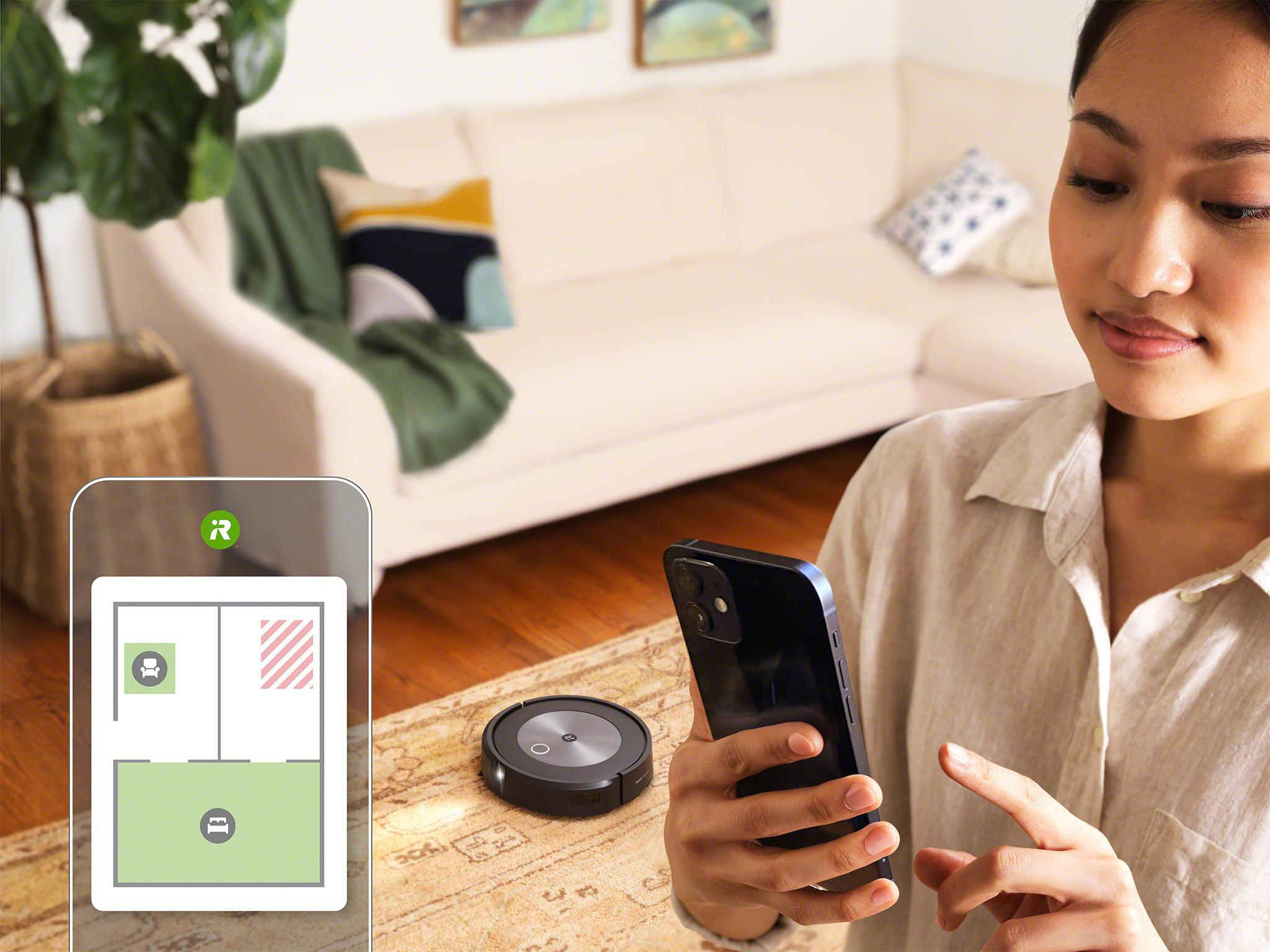 Frau benutzt Telefon neben Roomba-Roboter, zeigt Grundriss. Sofa im Hintergrund.