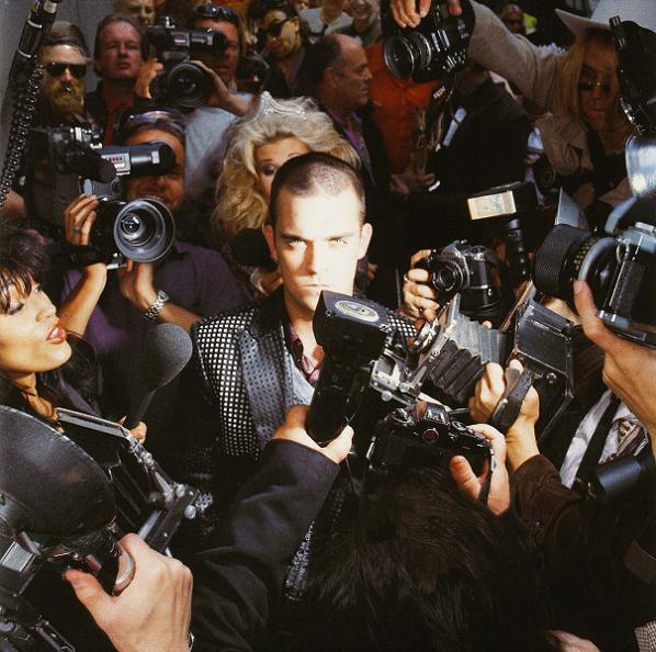 Ein Mann, umgeben von Paparazzi. Kameras und Mikrofone werden auf ihn gerichtet.