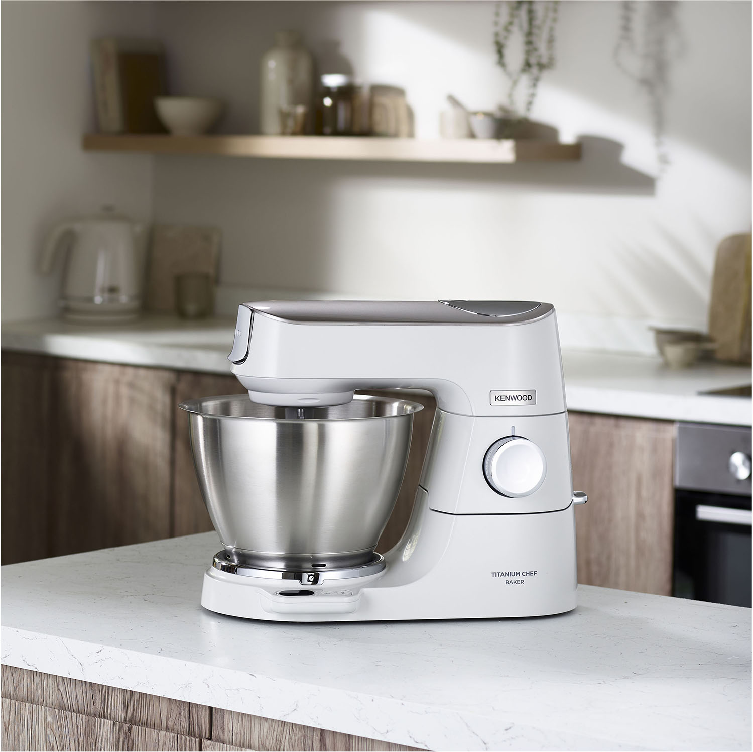 Een witte KitchenAid keukenmixer op een aanrecht. Achtergrond: houten kasten en een plank.
