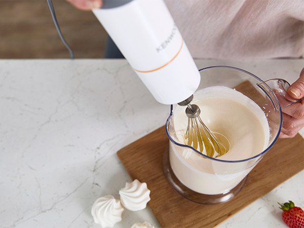 Hand holding a white Kenwood hand blender mixing cream in a clear pitcher.