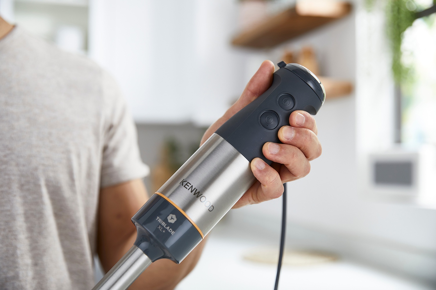 Hand die een zilveren en grijze Kenwood Triblade XL+ staafmixer vasthoudt. De hand staat in focus.
