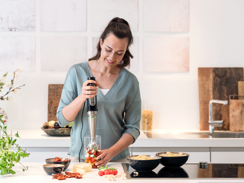 A woman using a hand blender, blending ingredients in a clear glass.