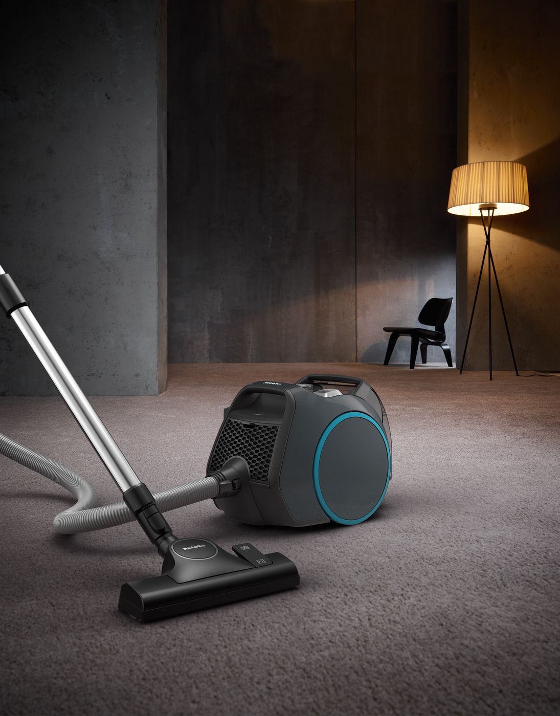 A grey vacuum cleaner with a blue ring sits on a grey carpet next to a lamp and chair.