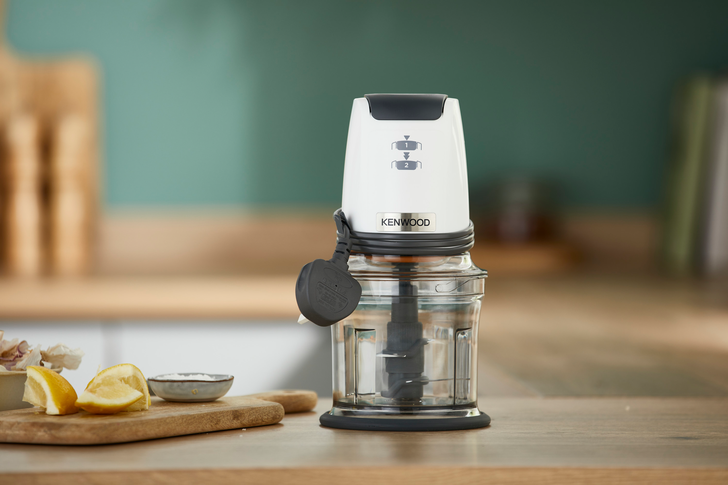 Weißer Kenwood-Zerkleinerer mit Glasschüssel und Holzbrett mit Zitronen.