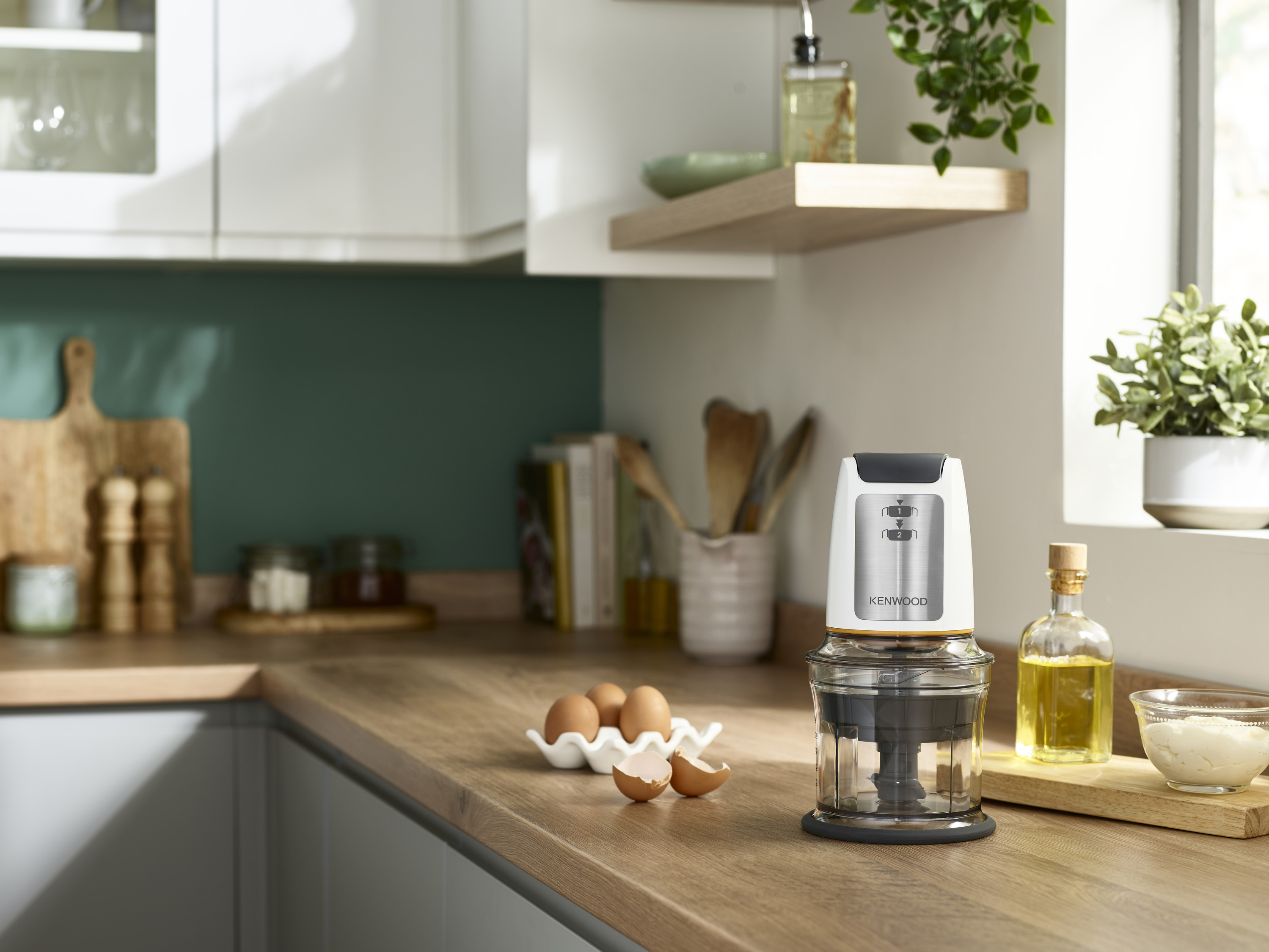 Ein Kenwood-Zerkleinerer auf einer Theke, mit Eiern und Olivenöl im Vordergrund. Küchenumgebung.