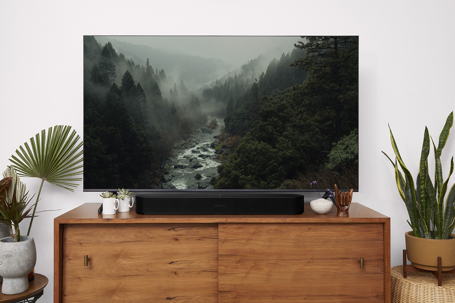 Een tv boven een houten kast toont een landschap. Een soundbar is onder de tv geplaatst. Planten staan aan weerszijden.