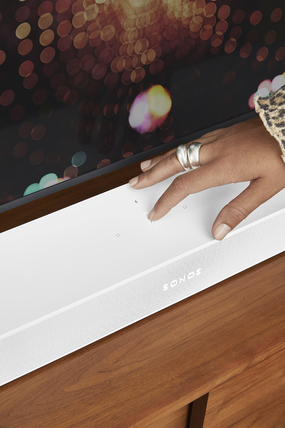 Een hand raakt de bovenkant van een witte soundbar op een houten oppervlak. Vage achtergrond met lichten.
