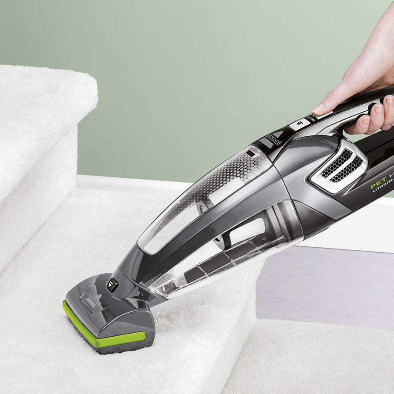 Handstaubsauger reinigt eine weiße, mit Teppich ausgelegte Treppe. Der Reiniger ist schwarz und grau.