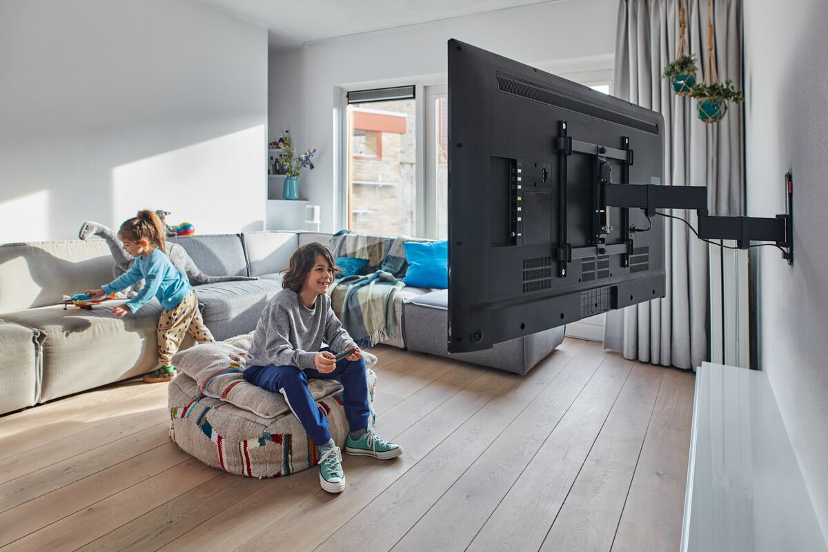 A boy on a beanbag chair plays a game on TV, another child plays behind him.