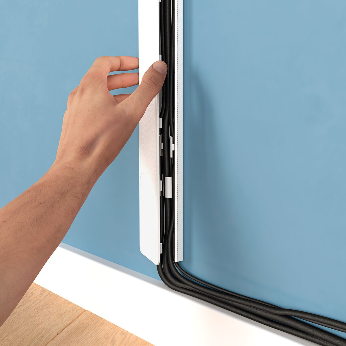 A hand installing a cable channel on a blue wall, with black cables.