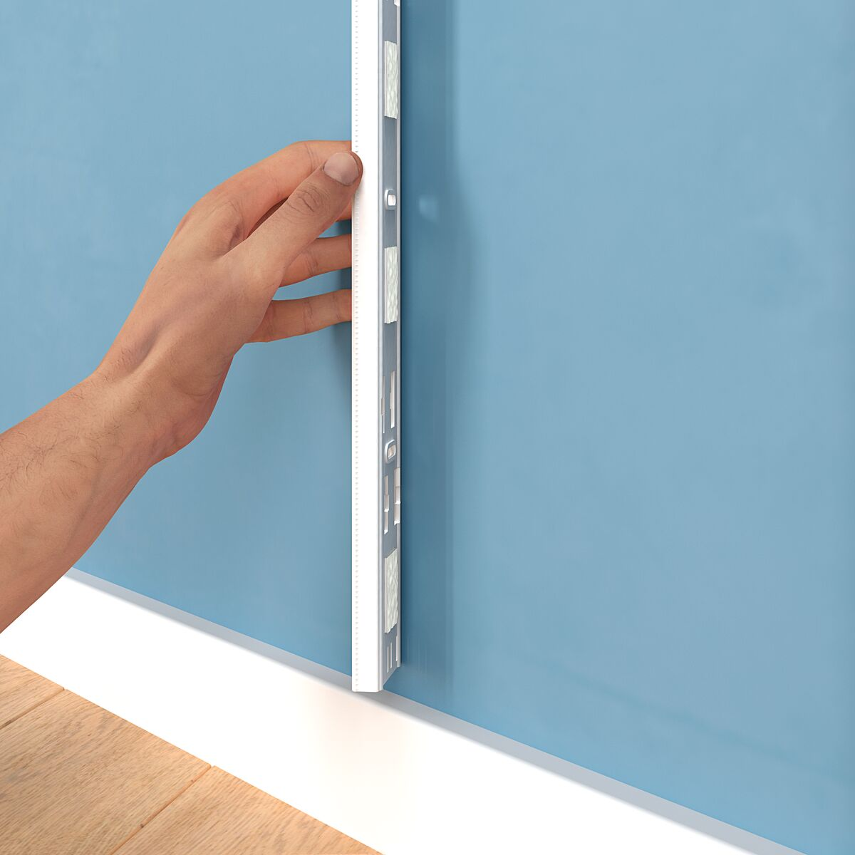 A hand holding a long, silver metal edge against a light blue wall. Wooden floor visible at the bottom.