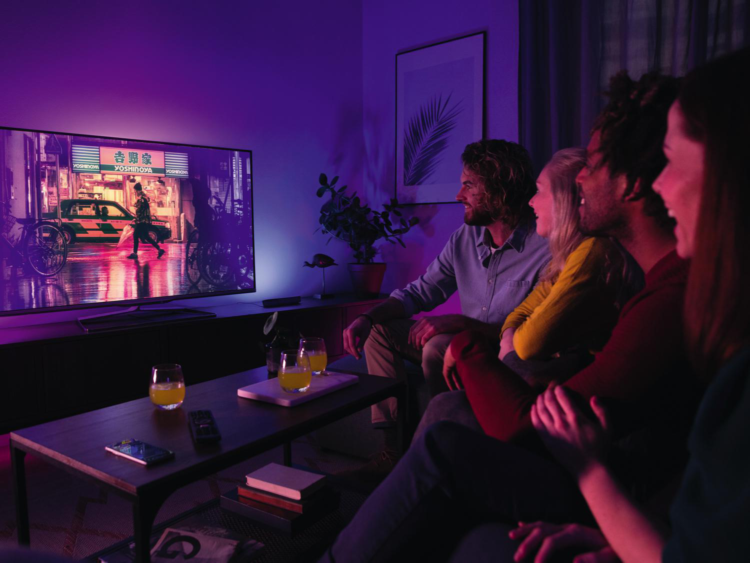 Leute schauen fern. Der Fernseher zeigt eine Stadtszene bei Nacht. Der Raum ist lila beleuchtet.