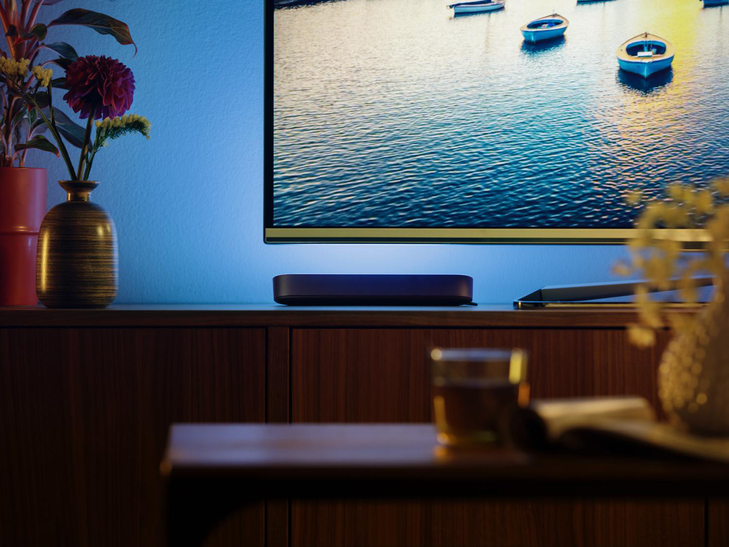 Ein Fernseher auf einem braunen Schrank, mit einer Soundbar darunter und einem Tisch mit einem Glas.