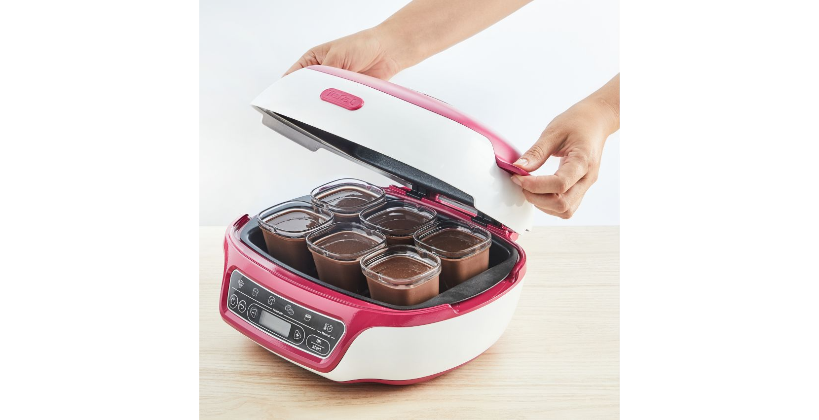 Een roze en witte yoghurtmaker met zes glazen potten, geopend door handen.