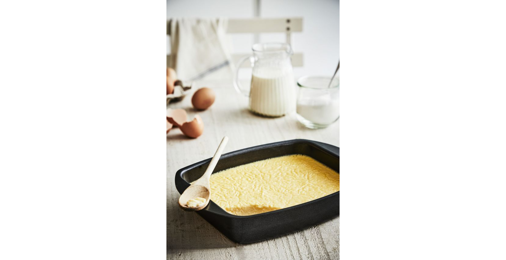 Een bakvorm met een geel, custardachtig mengsel, een houten lepel en ingrediënten op de achtergrond.