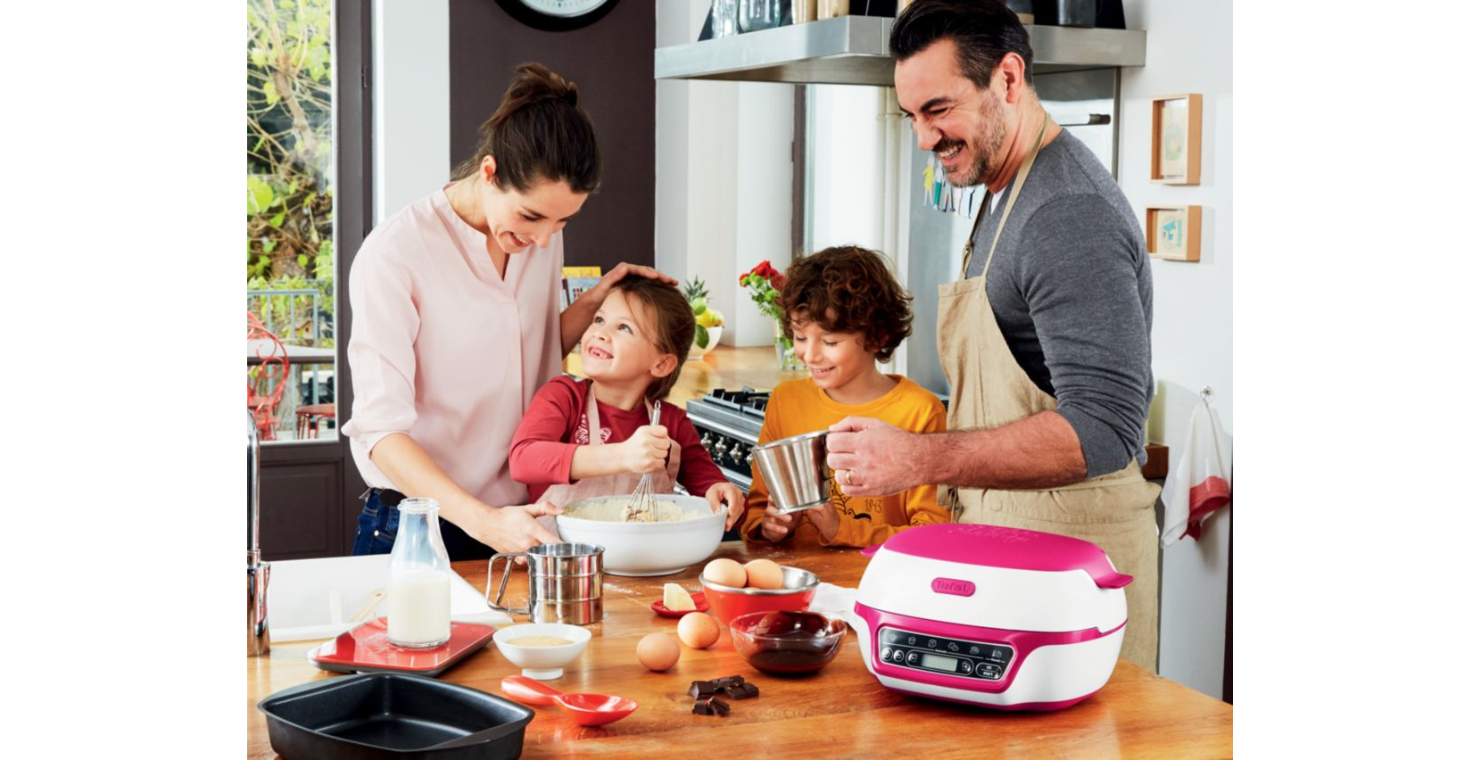 Familie bakt in een keuken. Vrouw en kinderen met ingrediënten en een roze apparaat op een tafel.