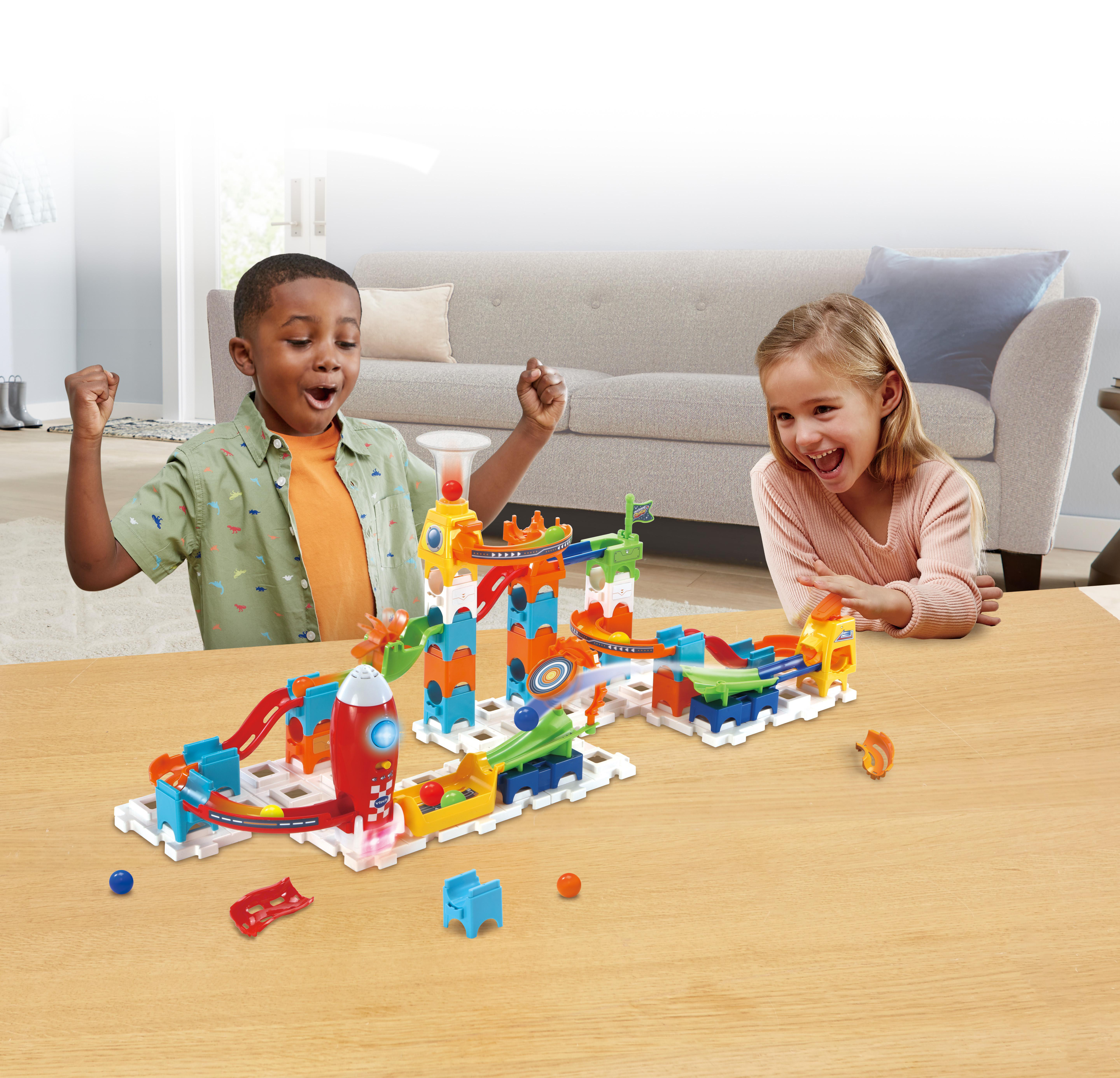 Zwei Kinder spielen mit einem bunten Murmelbahnspiel auf einem Tisch. Sie wirken aufgeregt und glücklich.