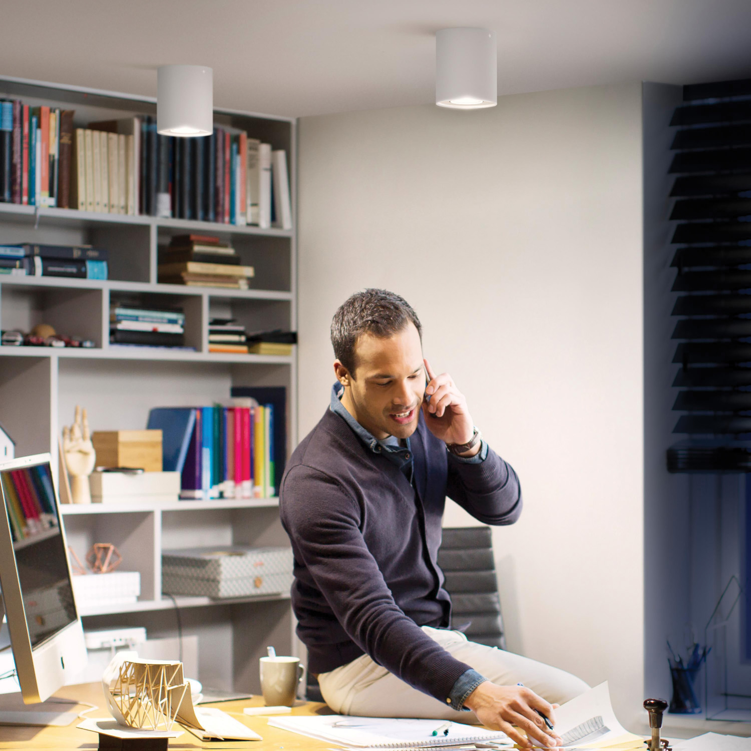 Homme sur un bureau au bureau, parlant au téléphone, papiers et étagère visibles.