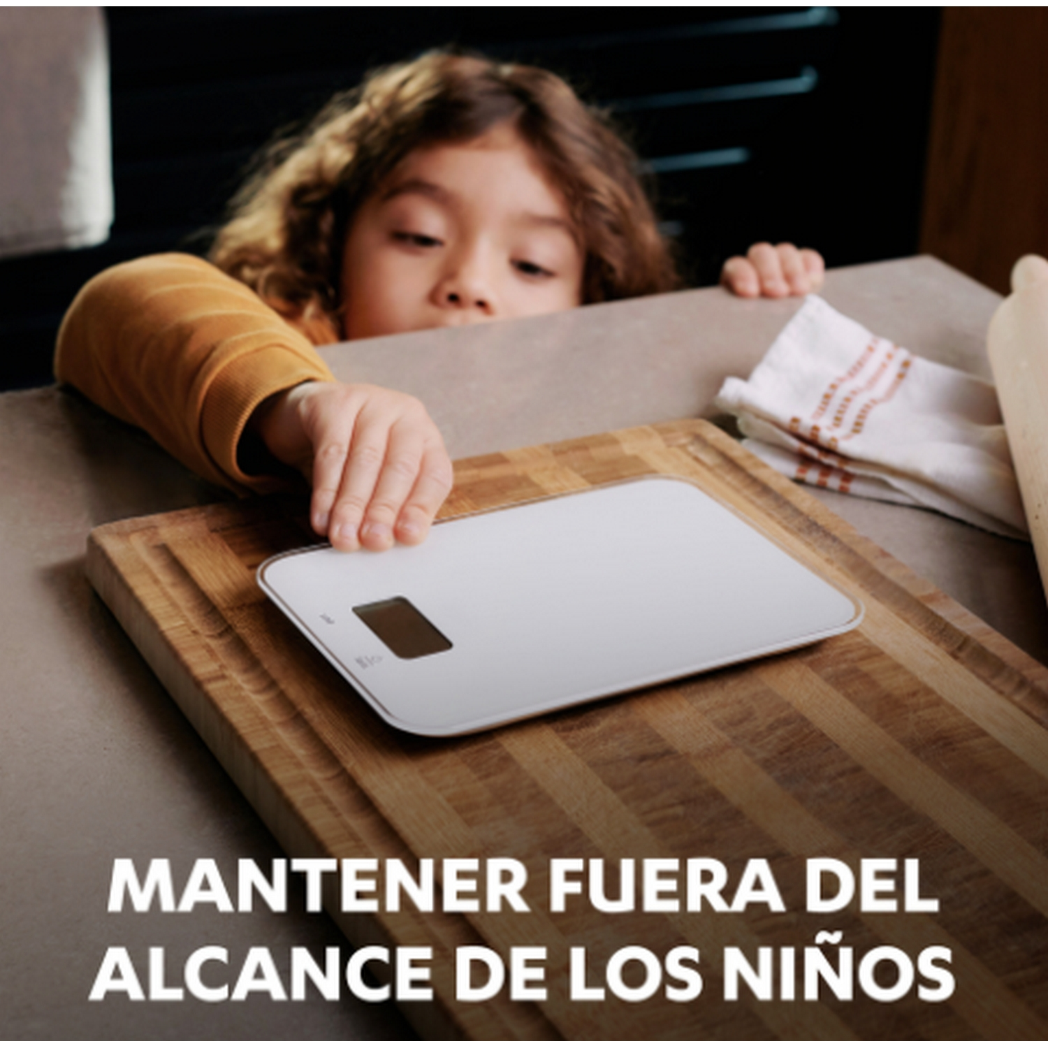 Manos de un niño alcanzan una balanza blanca sobre una tabla de cortar de madera, con texto debajo.
