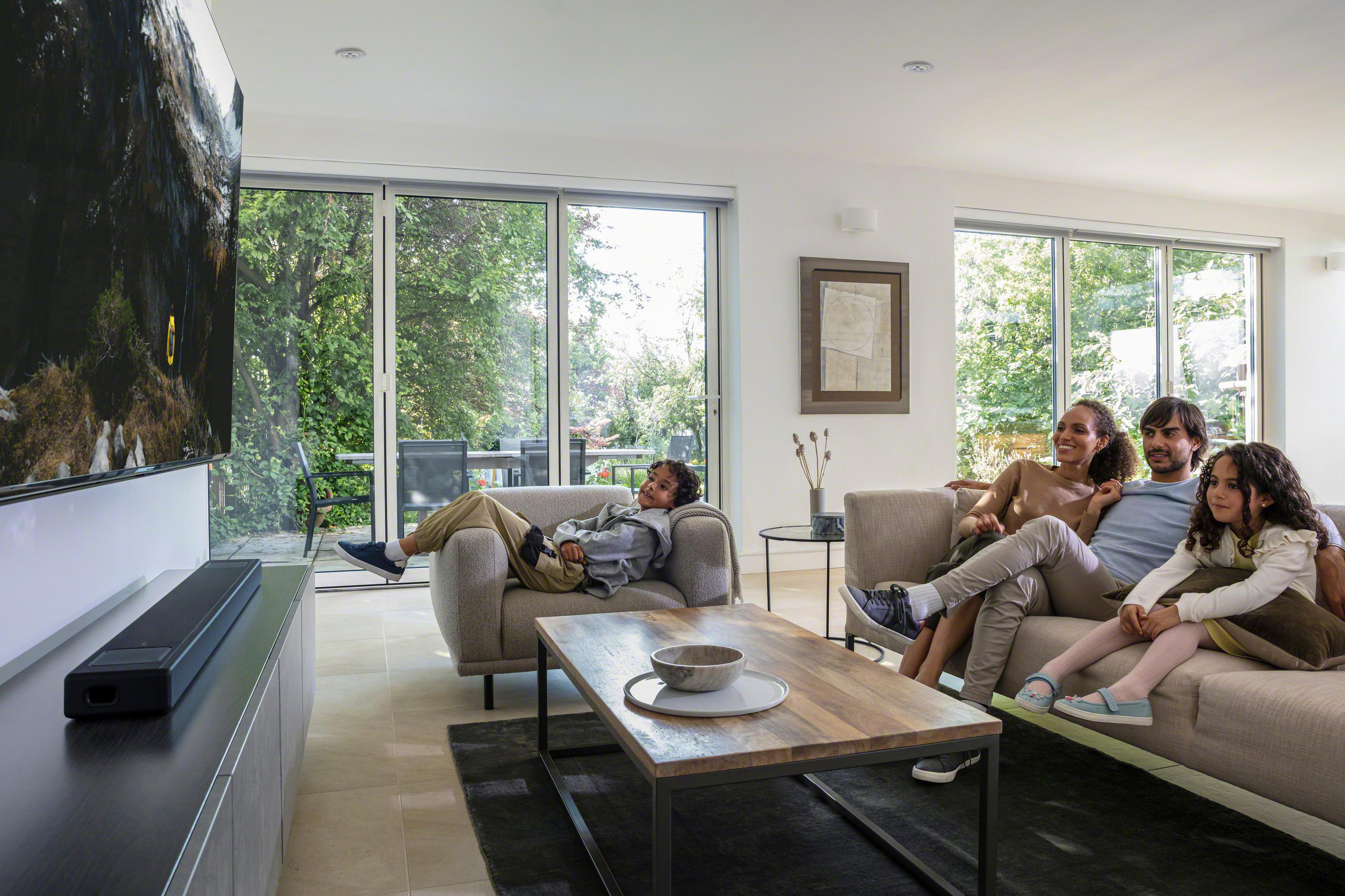 Familie im Wohnzimmer, beim Fernsehen. Holztisch und Sofa vor einem großen Fenster.