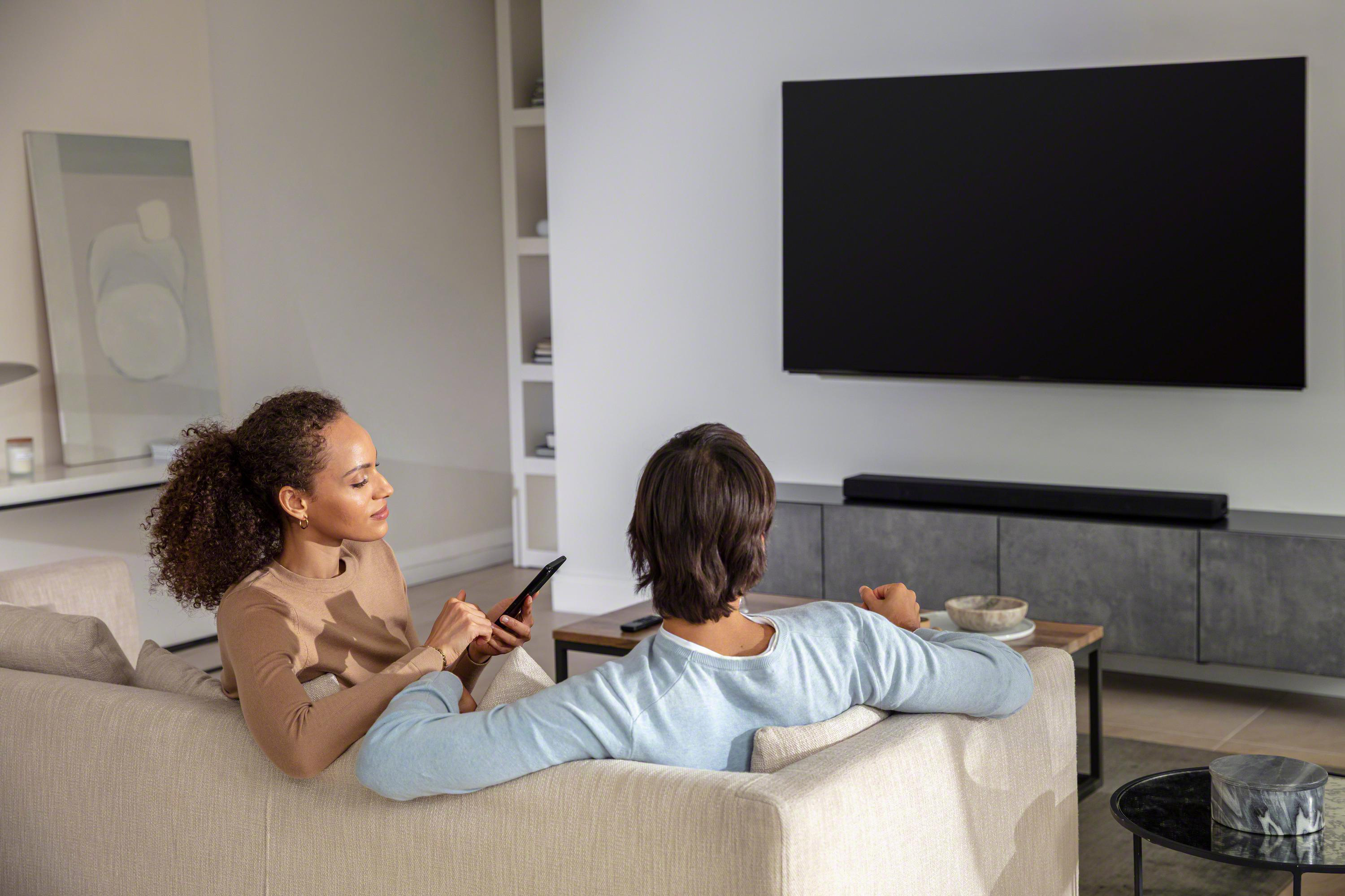 Paar auf einem Sofa, das auf einen Fernsehbildschirm schaut. Eine Soundbar unter dem Fernseher.