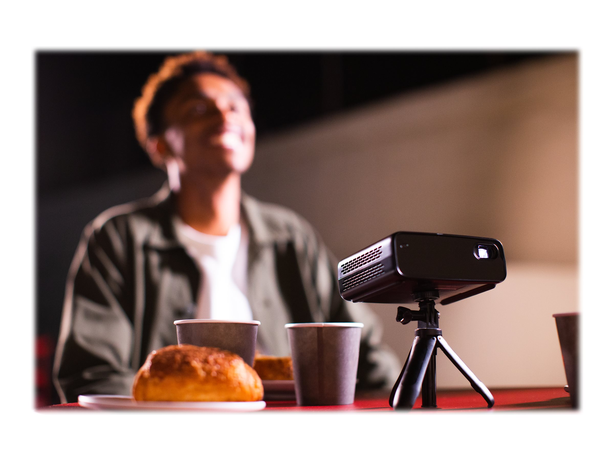 Zwarte projector op een statief, met eten en een persoon. Rode tafel, nacht.