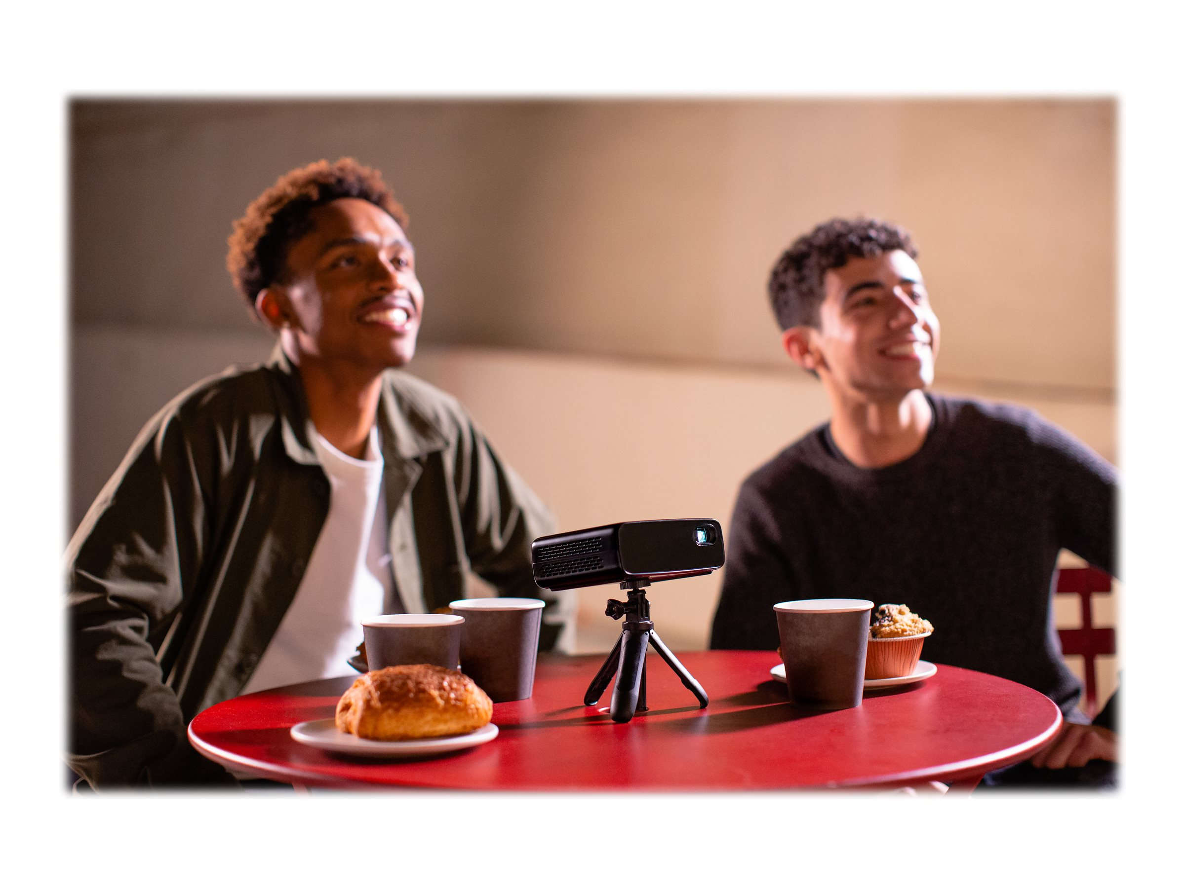 Twee mannen kijken naar een kleine projector op een statief, met snacks op een rode tafel.