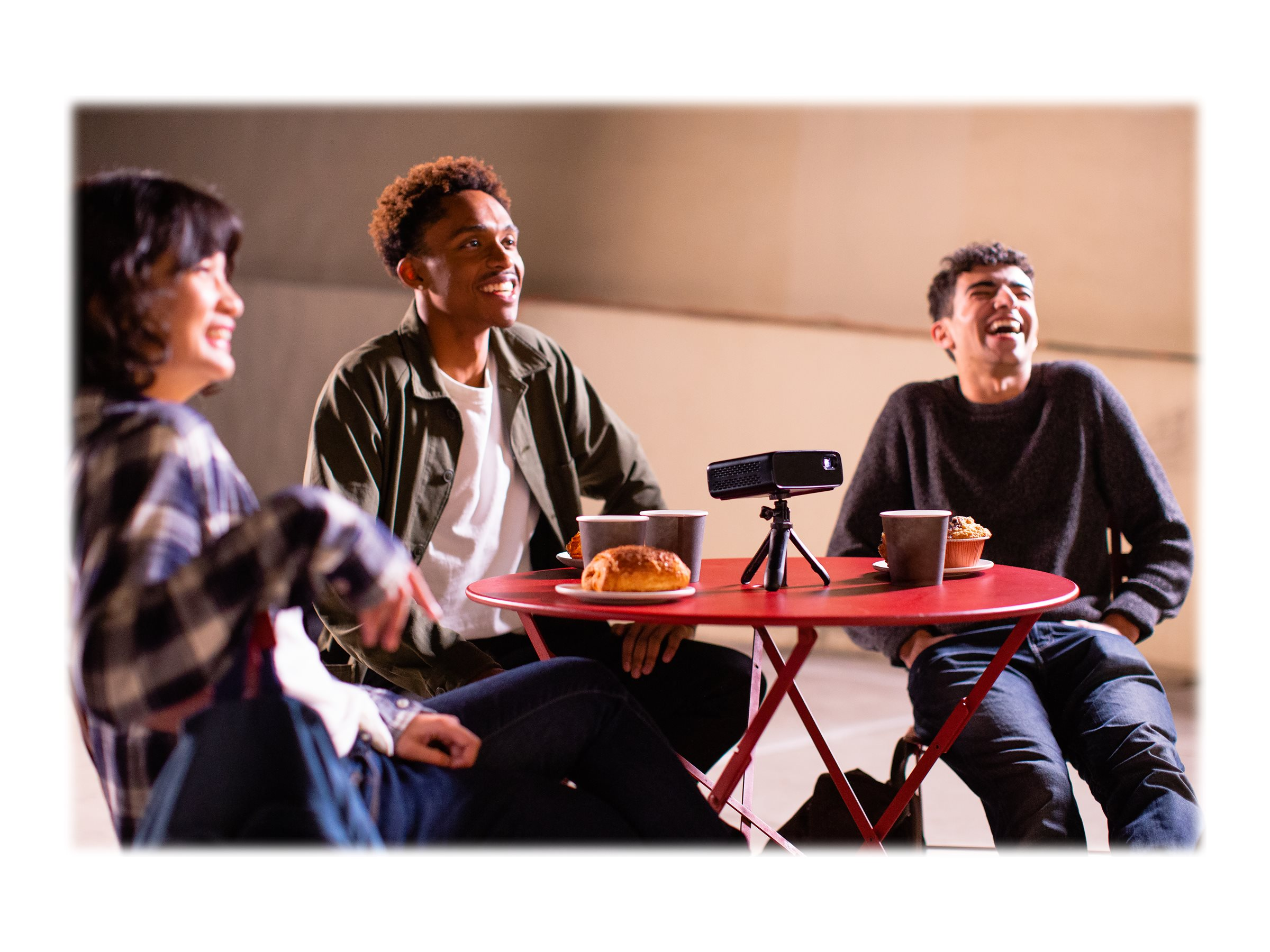 Drie mensen lachen rond een rode tafel met een projector en eten.