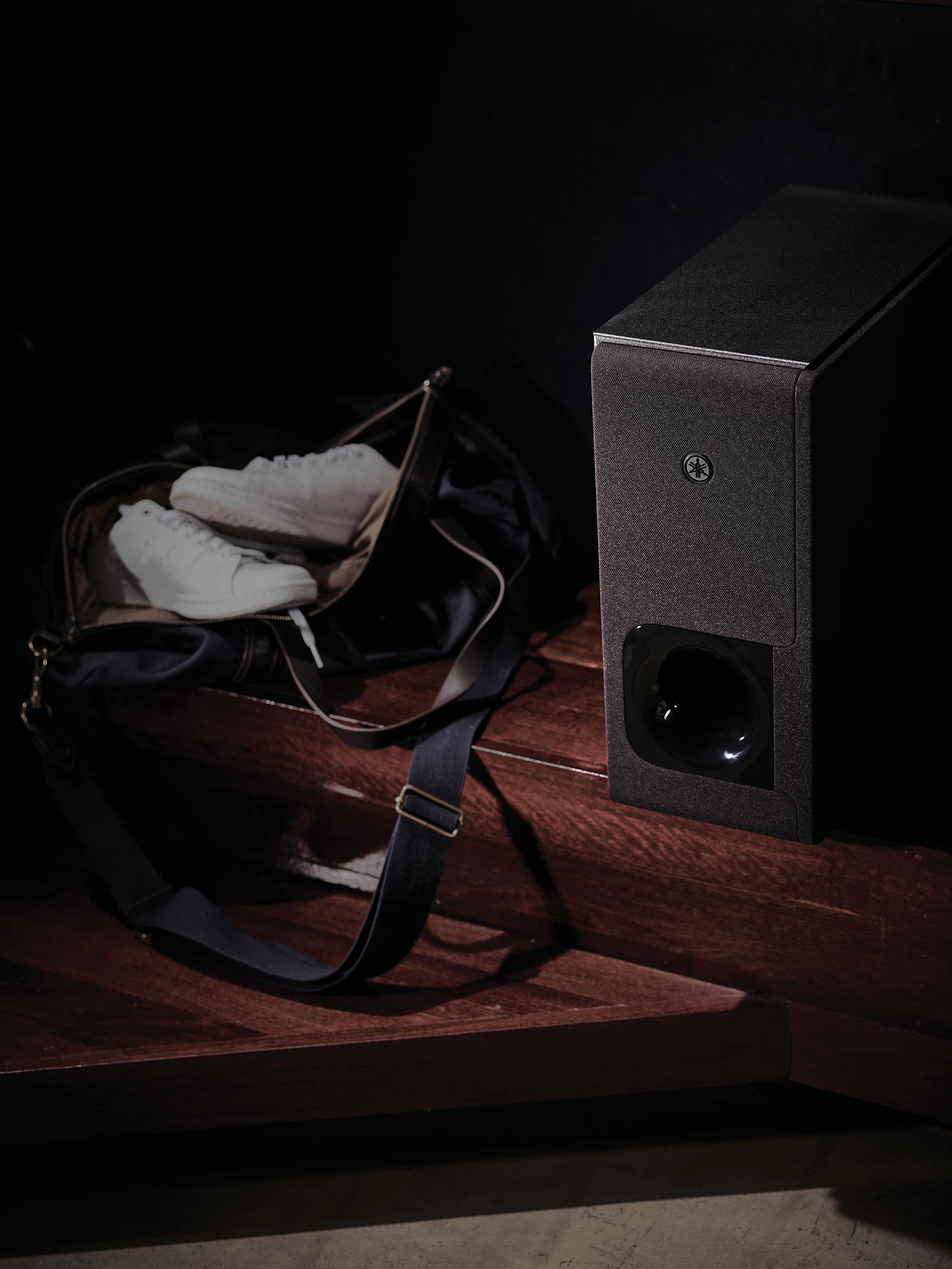 Ein schwarzer Lautsprecher neben einer schwarzen Tasche mit weißen Turnschuhen. Dunkle Holzoberfläche.