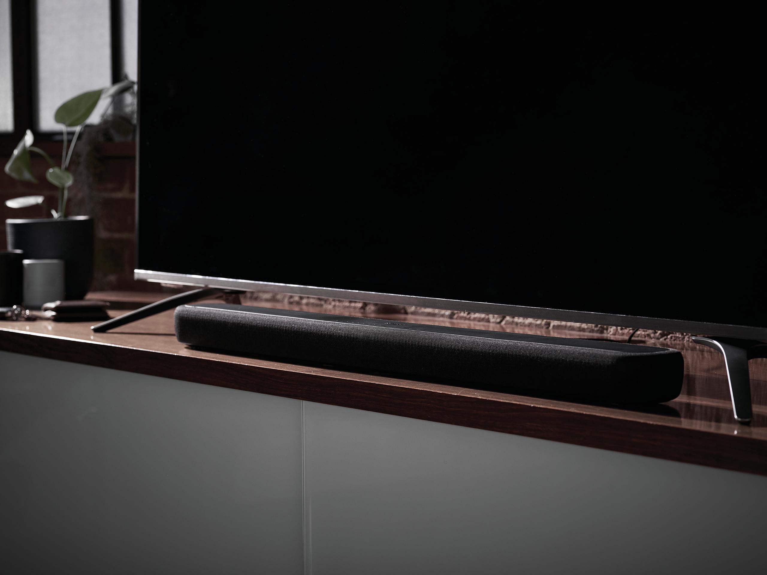 Eine schwarze Soundbar auf einem Holzregal unter einem Fernseher vor einer Ziegelmauer.