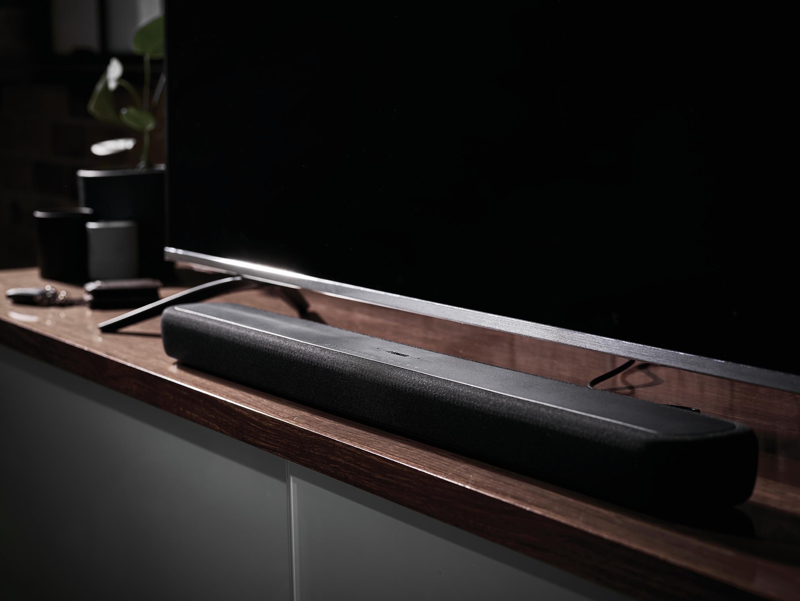 Schwarze Soundbar auf Holzoberfläche neben einem Fernseher.
