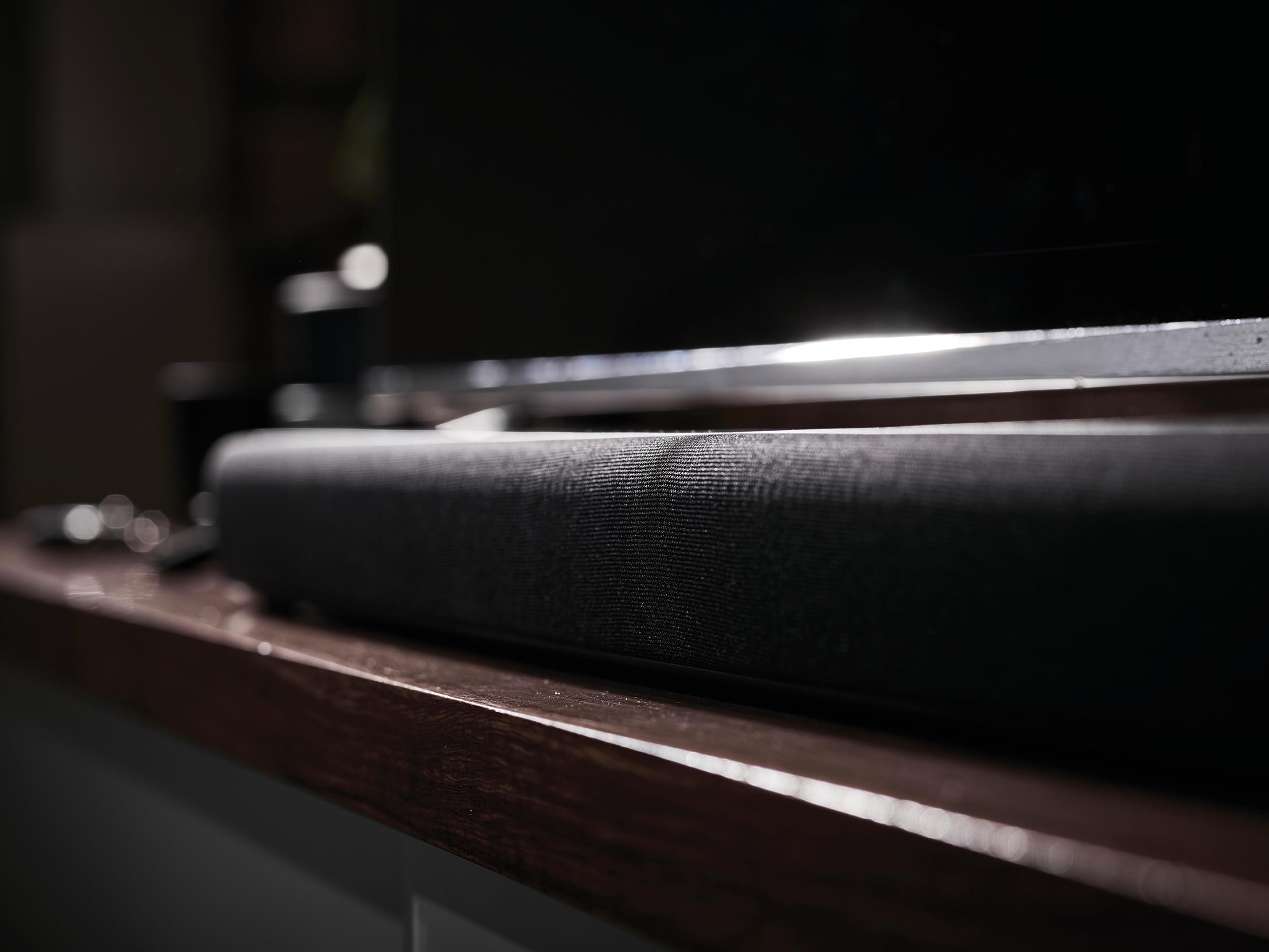 Eine schwarze Soundbar auf einer Holzoberfläche. Die Soundbar ist im Fokus und befindet sich in der Nähe eines Fernsehers.