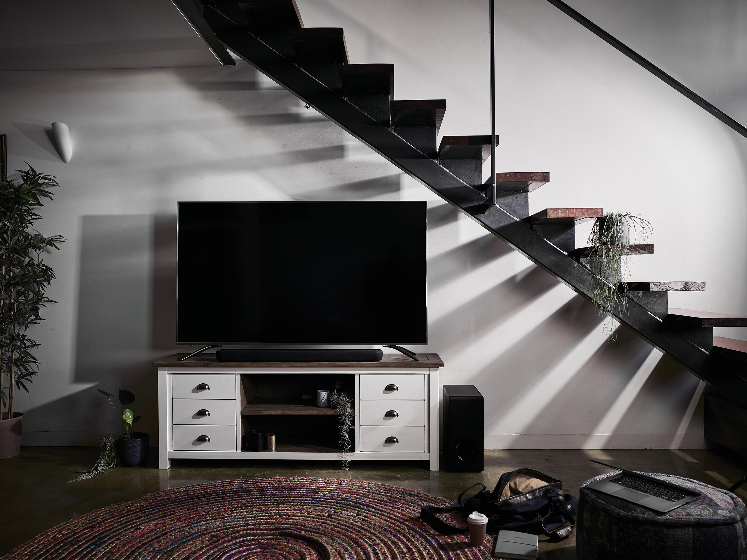 Wohnzimmer mit Fernseher auf weißem Schrank. Treppe rechts. Teppich, Lautsprecher, Pflanzen und Laptop.