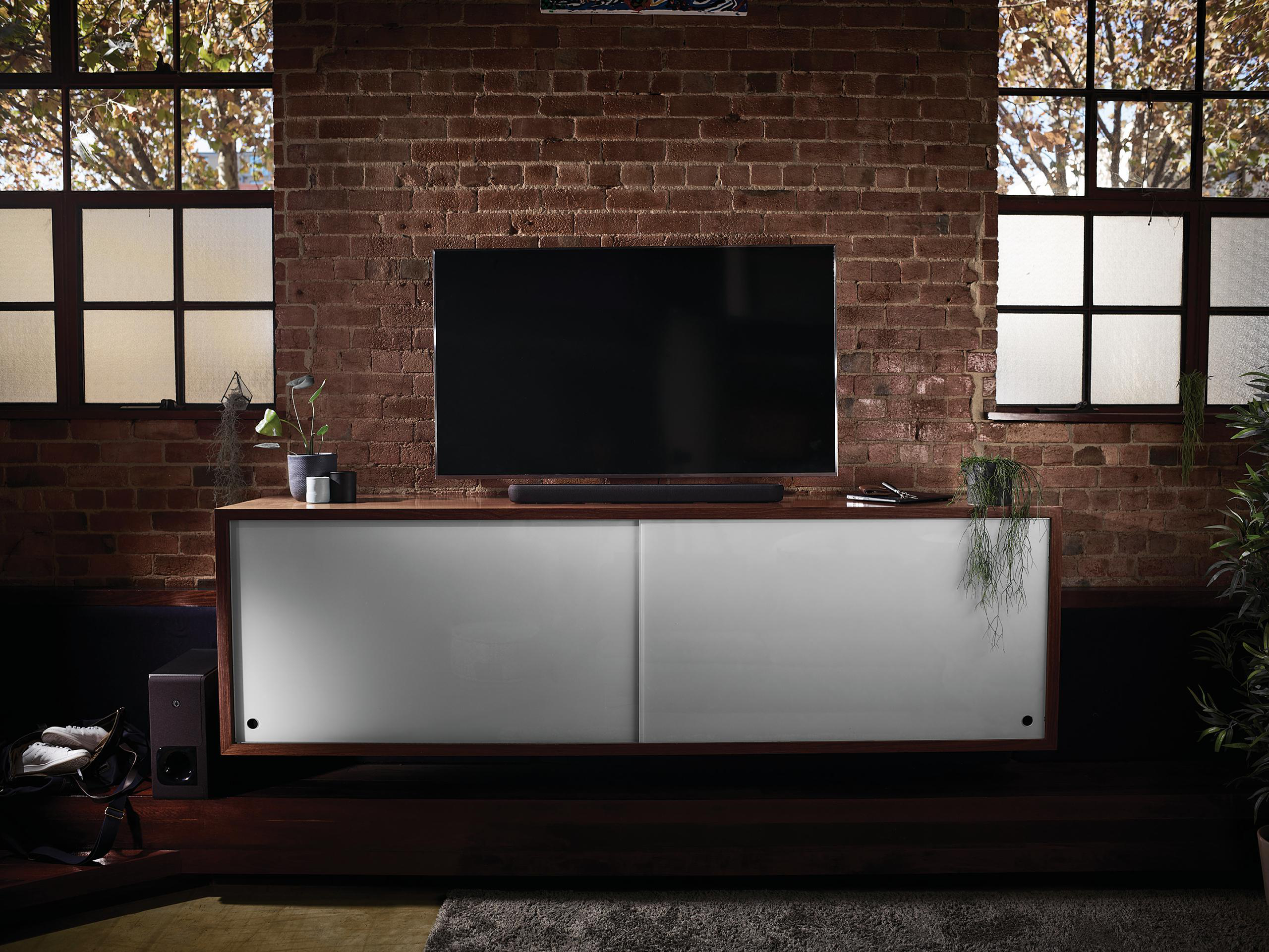 Modernes Wohnzimmer mit großem Fernseher, Soundbar und weißem Schrank. Backsteinwand im Hintergrund.