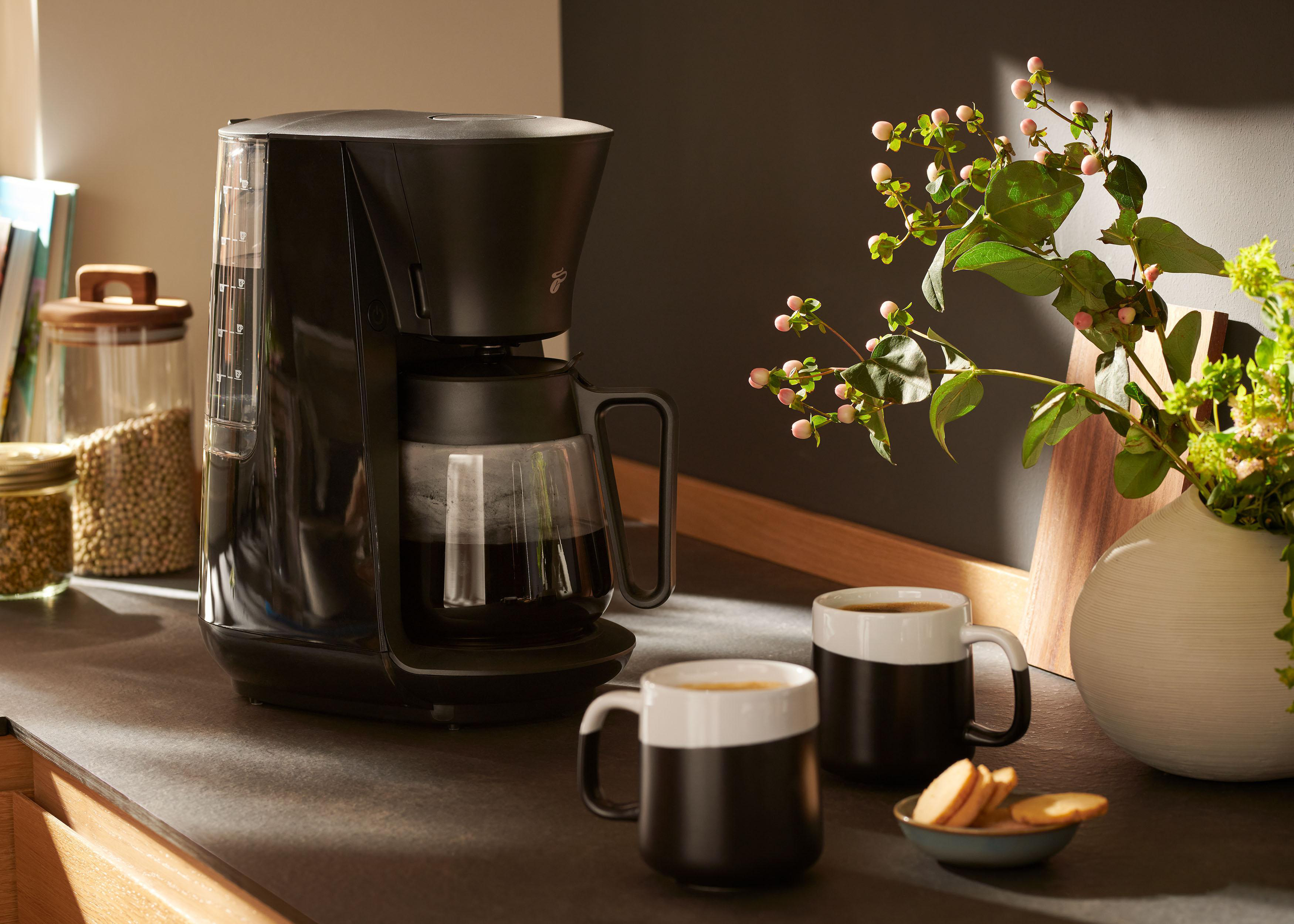 Schwarze Kaffeemaschine mit Glaskaraffe, zwei Tassen und einem Teller Keksen, neben Blumen.