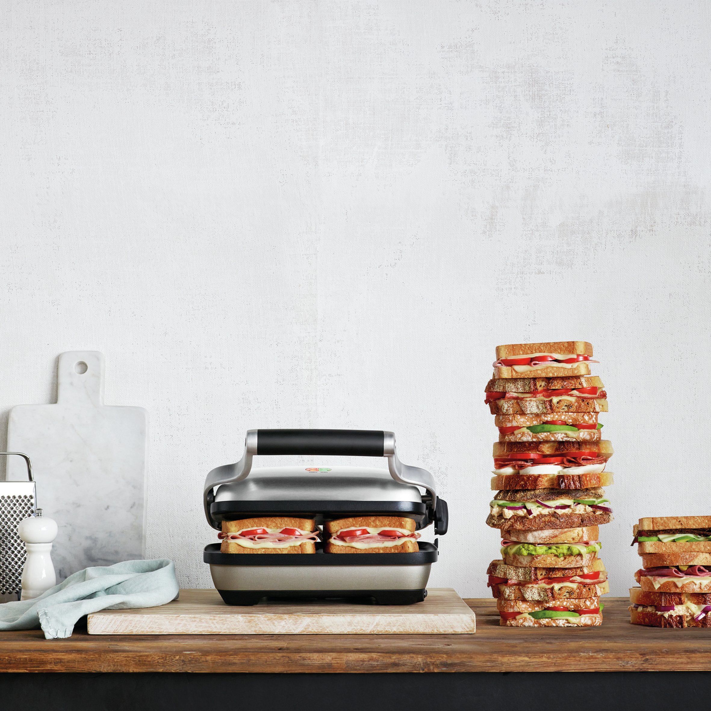 Sandwichmaker, mit zwei Sandwiches darin und Sandwichstapeln auf einem Holztisch.