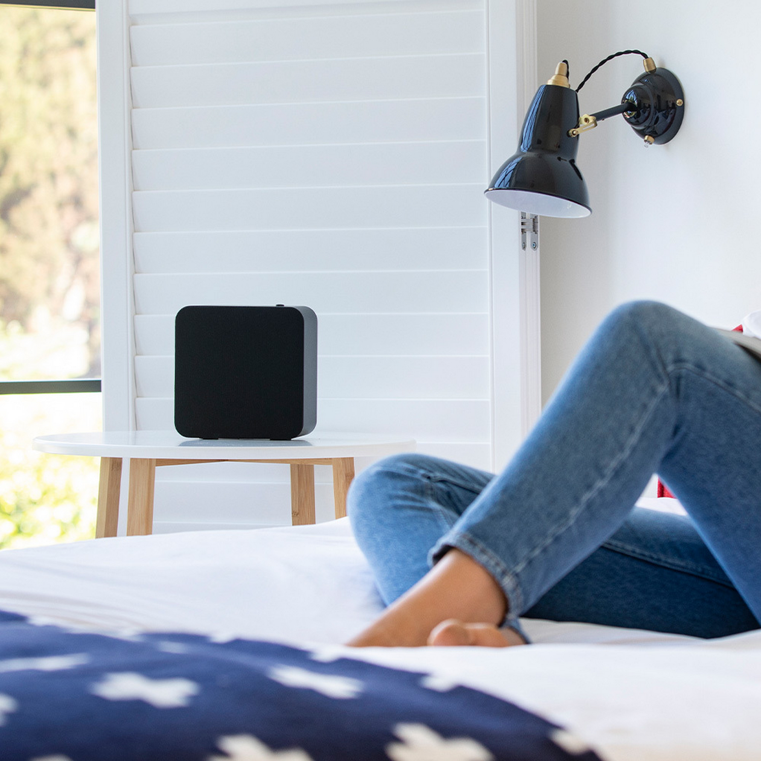 Una persona con jeans se relaja en una cama junto a un altavoz negro sobre una mesa cerca de una ventana.