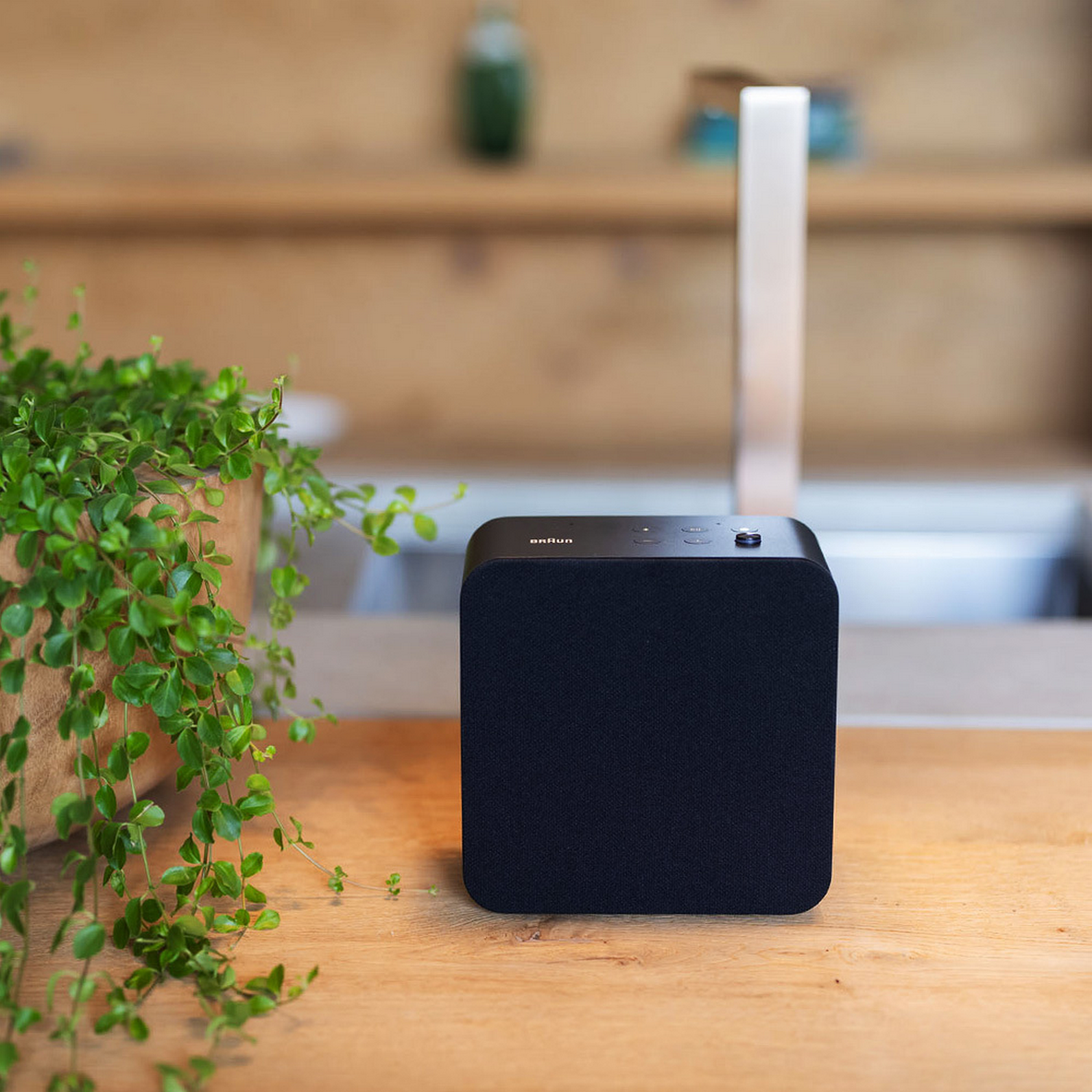 Un altavoz negro sobre una mesa de madera, plantas verdes a la izquierda y una cocina borrosa en el fondo.