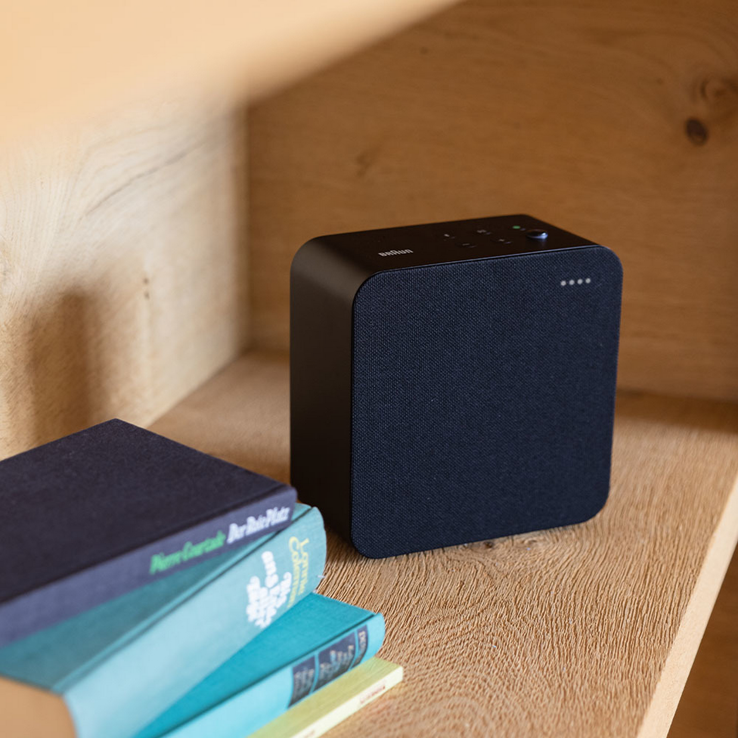 Altavoz cuadrado negro en estante de madera con libros en primer plano. Diseño simple y minimalista.