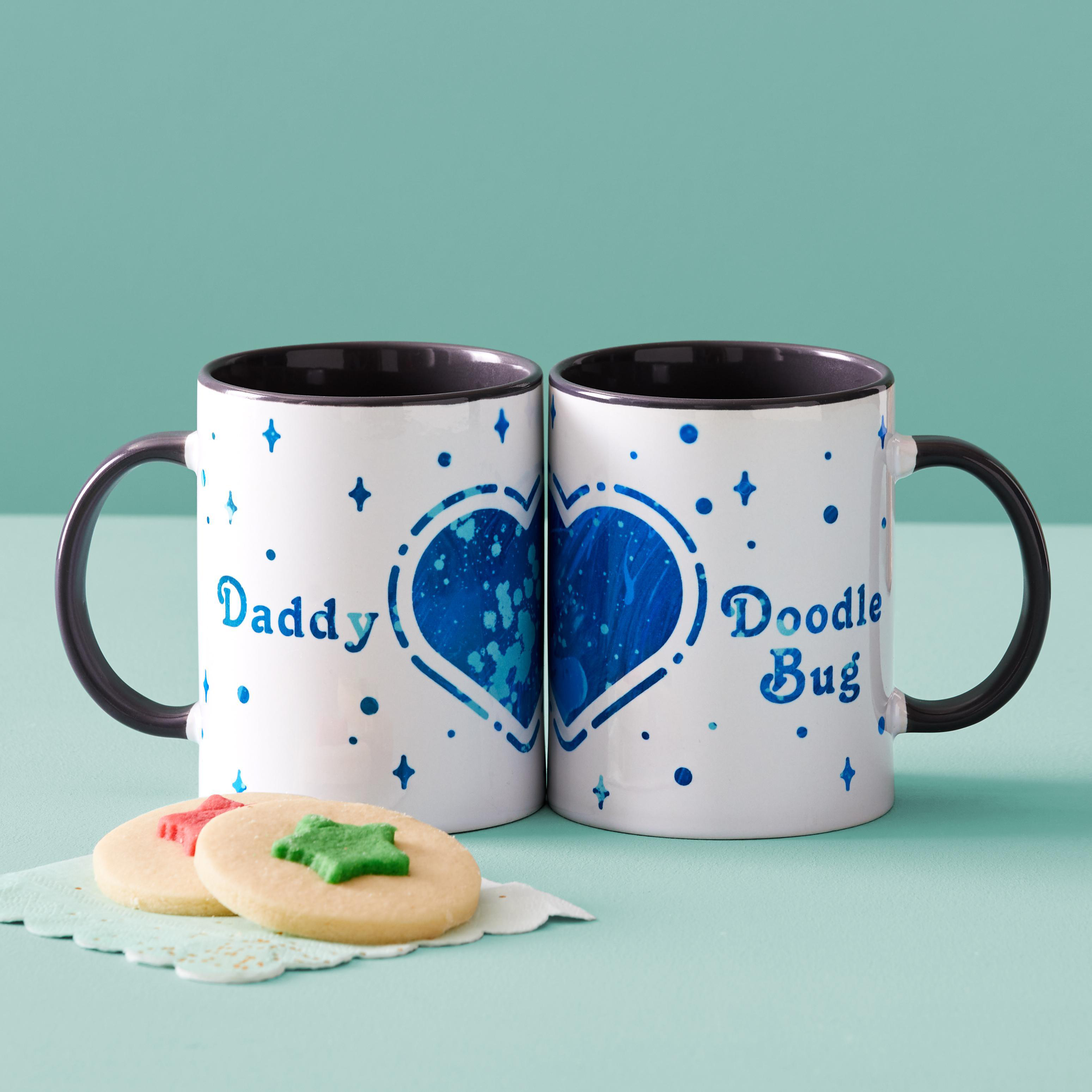 Zwei weiße Tassen mit schwarzen Griffen. Eine sagt 'Daddy', die andere 'Doodle Bug', mit Keksen davor.