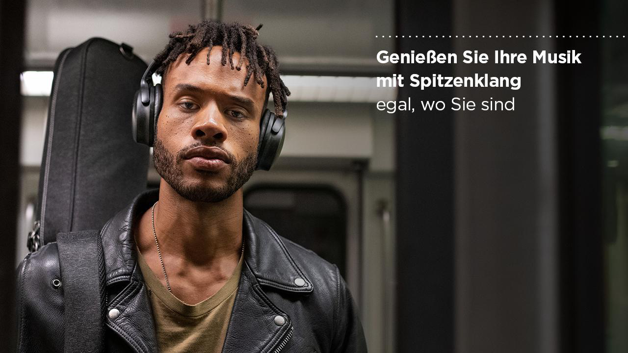 Ein Mann in einer schwarzen Lederjacke trägt Kopfhörer in der U-Bahn. Eine Gitarrentasche.