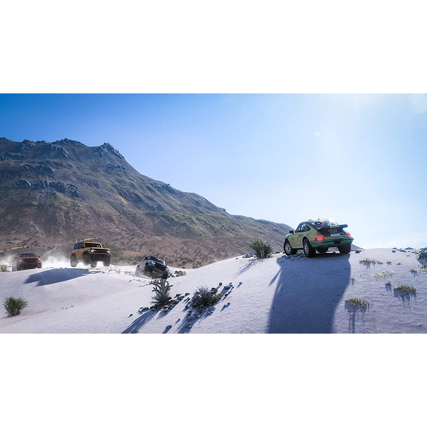 Coches conduciendo en un camino arenoso con montañas al fondo, cielo azul.