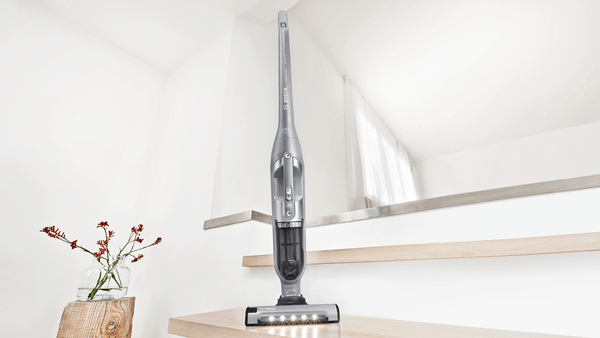 Grauer Stiel-Staubsauger steht auf einer Holzstufe. Eine Blumenvase steht daneben.