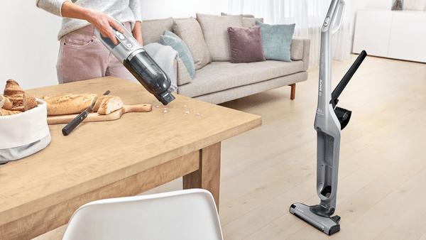 Person mit einem kabellosen Staubsauger auf einem Holztisch, Sofa im Hintergrund.