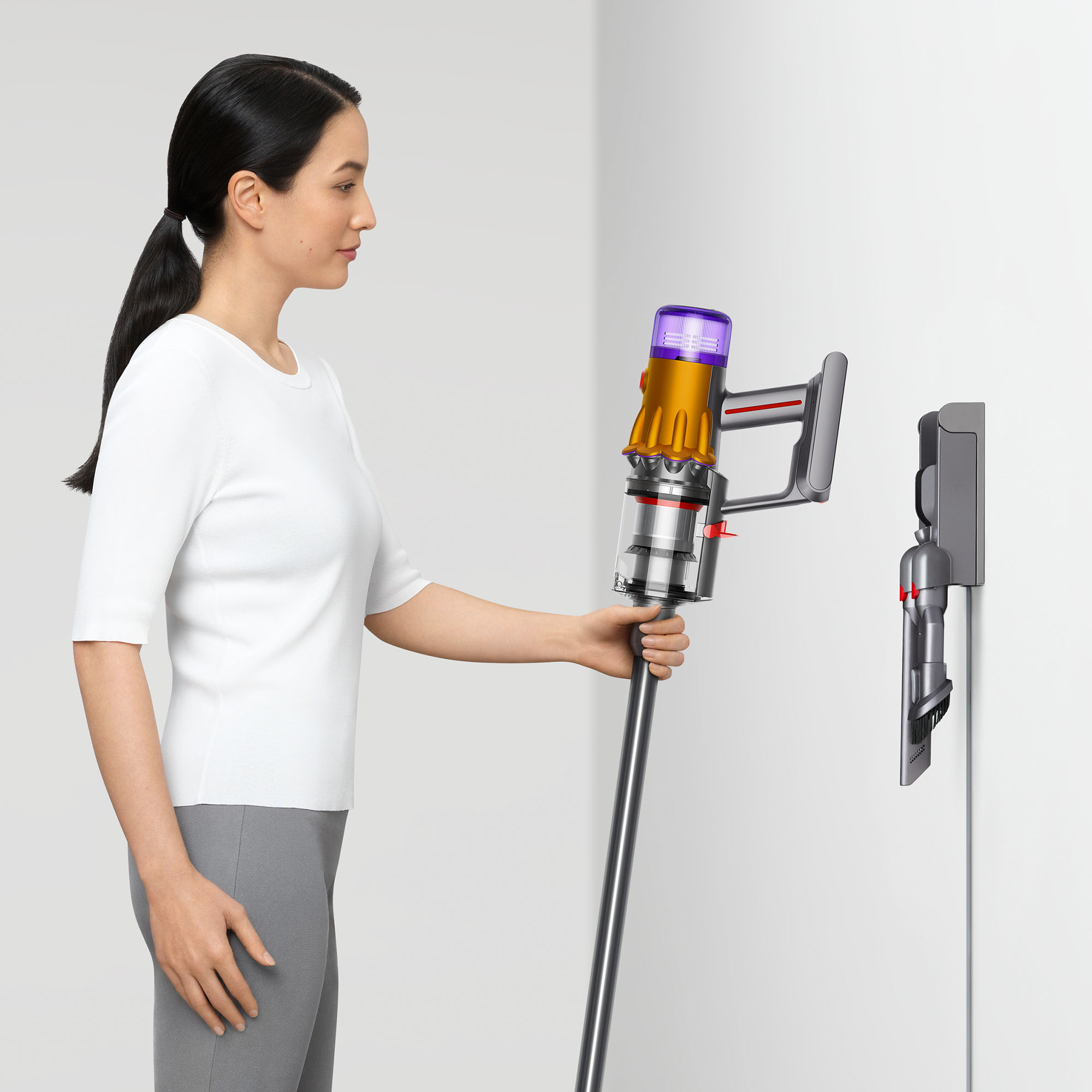Beyaz gömlekli kadın kablosuz elektrik süpürgesi kullanıyor. Elektrik süpürgesi arka planda duvara monte edilmiş.