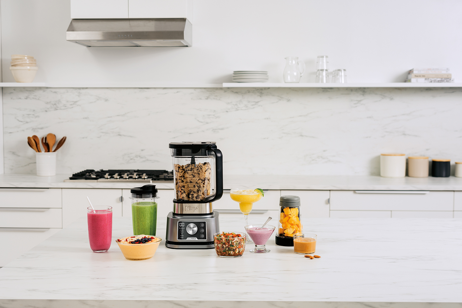 Een blender met verschillende smoothies en ingrediënten op een wit aanrecht in een keuken.