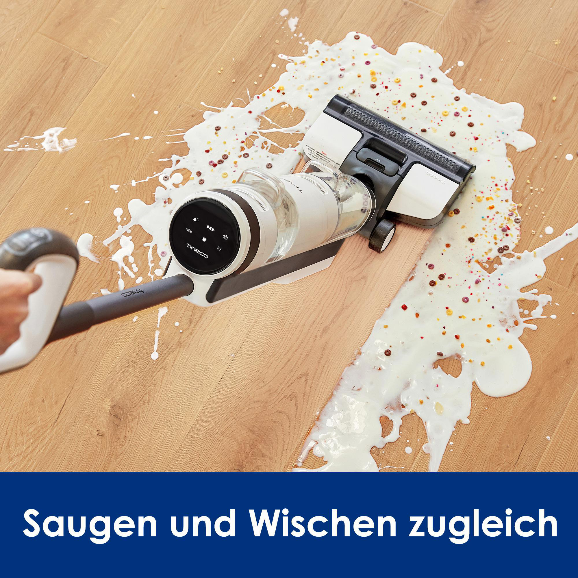 Staubsauger reinigt verschüttete Milch und Müsli auf einem Holzboden. Der Staubsauger ist weiß und grau.
