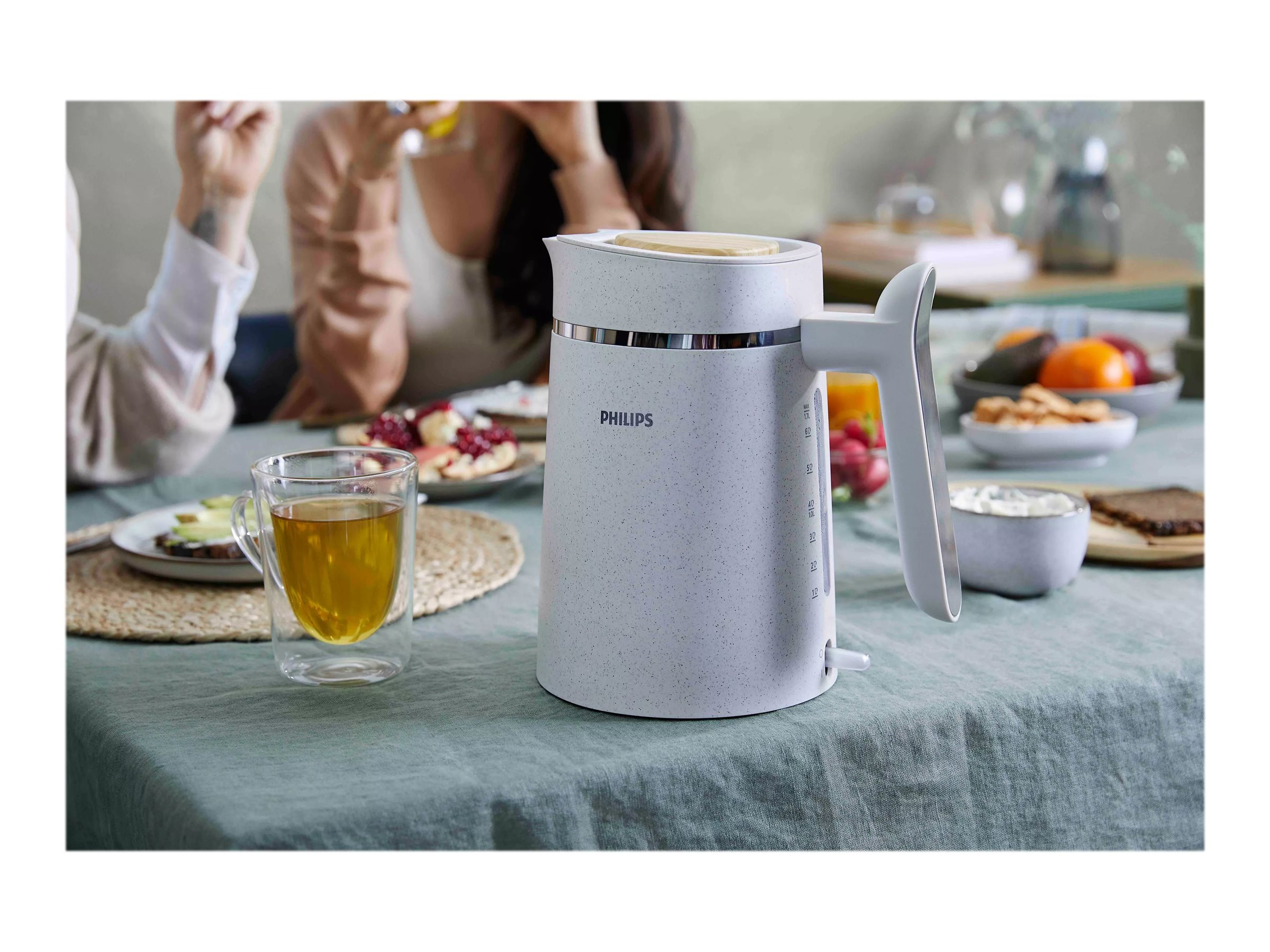 Ein weißer Wasserkocher auf einem Tisch mit Essen und einer Teetasse. Personen im Hintergrund.
